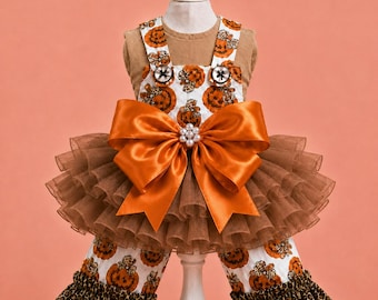 Puede incluir: Una camiseta color canela con un peto blanco con estampado de calabazas naranjas y leopardo. El peto tiene un gran lazo de satén naranja con un centro de perlas, volantes de tul marrón en la cintura y bajos de pantalón con volantes de leopardo.