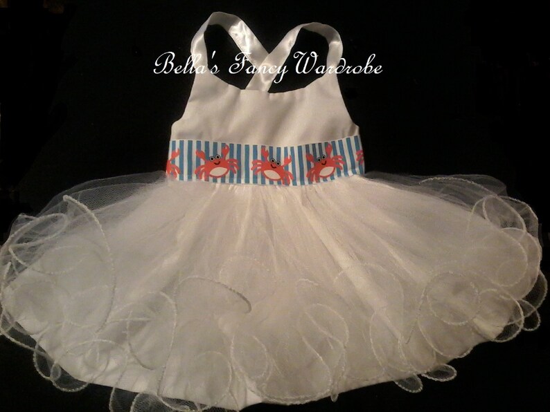 May include: White tulle dress with a white satin bodice and a blue and white striped ribbon with red crab appliques. The dress has a ruffled hem.