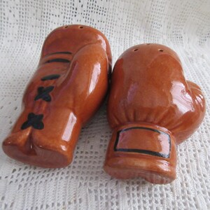 Puede incluir: Un par de saleros y pimenteros de cerámica marrón con forma de guantes de boxeo. Los guantes tienen detalles negros y son brillantes. Los saleros y pimenteros están sobre una superficie blanca texturizada.