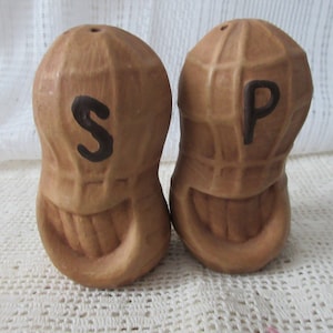 May include: A set of ceramic peanut salt and pepper shakers. The shakers are brown and have a peanut shell design. The salt shaker has the letter "S" on it and the pepper shaker has the letter "P" on it.