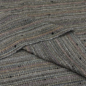 Op de afbeelding: Close-up van een textiel met een geweven patroon. De stof heeft een mix van kleuren, waaronder bruin, grijs en hints van paars en goud. De weving creëert een enigszins ruw, tactiel oppervlak, geschikt voor kleding of woondecoratie.