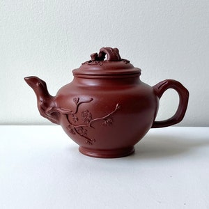 May include: A reddish-brown ceramic teapot with a curved spout, handle, and lid. The teapot is decorated with floral and branch designs. The lid has a decorative handle. The teapot is sitting on a white surface.