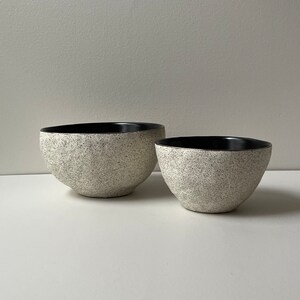 May include: Two speckled ceramic bowls with black interiors. The bowls have a rounded shape and a textured, speckled exterior in shades of white and gray. The bowls are of different sizes and are placed side by side.