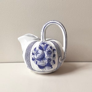 May include: White ceramic pitcher with a blue floral design. The pitcher has a curved handle and a spout. The floral design is on the front and sides of the pitcher. The handle is decorated with blue lines.