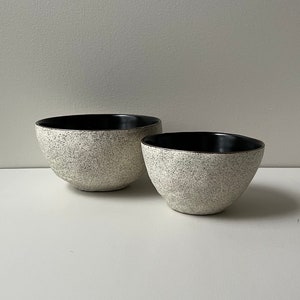 May include: Two ceramic bowls. The bowls have a speckled, off-white exterior and a black interior. The bowls are different sizes and have a rounded shape. The bowls are sitting on a white surface.