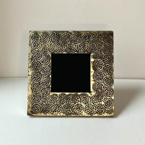 May include: Square picture frame with a black interior and a gold-colored, textured border. The border features a repeating spiral design. The frame is sitting on a white surface.