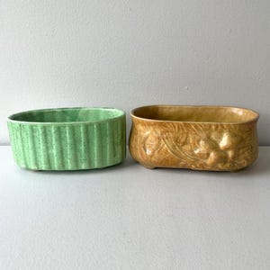 May include: Two ceramic planters. One is light green with vertical ribbed detailing. The other is a tan-colored planter with a floral relief design. Both are rectangular with rounded corners.