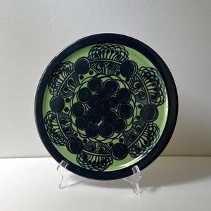 May include: Decorative ceramic plate with a black rim and a light green center. The center features a black floral design with circular and stylized elements. The plate is displayed on a clear plastic stand, showcasing its intricate pattern.
