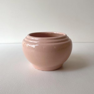 May include: A pale pink ceramic planter with a rounded shape and a smooth, glossy finish. The planter has three horizontal ridges around the top edge.