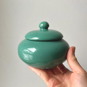 May include: A teal ceramic jar with a lid. The jar is round with a smooth, glossy finish. The lid has a small, rounded knob on top. The jar is held in a hand against a plain white background.