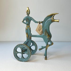 May include: A teal and gold-toned metal sculpture of a chariot with a figure and a horse. The figure is standing in the chariot, and the horse is rearing up. The wheels have a Greek key design. A tag is attached to the sculpture.