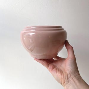 May include: A pale pink ceramic planter with a rounded shape and a slightly flared rim.
