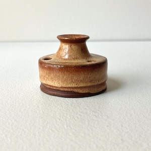 May include: A small, handcrafted ceramic inkwell with a brown and tan glaze. The inkwell has a cylindrical base and a flared top with a small opening. The piece has a rustic, vintage aesthetic.