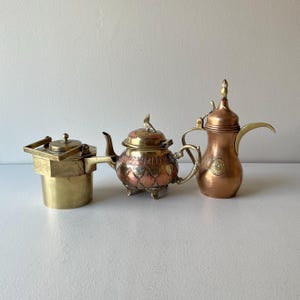 May include: Three antique brass and copper teapots. The teapot on the left is a small, round, brass teapot with a lid. The teapot in the center is a larger, round, brass and copper teapot with a lid and a decorative design. The teapot on the right is a tall, copper teapot with a lid and a spout.