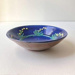 May include: A handmade ceramic bowl with a blue interior and a brown exterior. The blue interior is decorated with a green vine and yellow floral accents. The bowl is round and has a shallow depth.