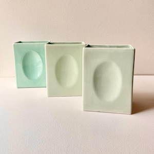 May include: Three ceramic vases in shades of green, each with a unique oval indentation. The vases are square in shape and have a smooth, matte finish.