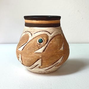 May include: A small, rounded ceramic pot with a textured tan and cream surface featuring carved abstract patterns. The rim is dark brown with a horizontal band, and a single small turquoise-colored stone is embedded in the center design.