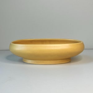 May include: A yellow ceramic planter with a speckled finish. The oval-shaped bowl sits on a low, circular base. The planter has a smooth, rounded rim and a slightly textured surface.