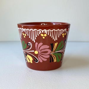May include: A small, terracotta-colored ceramic pot with hand-painted floral designs in pink, yellow, green, and black. The rim features a white and yellow pattern. This decorative pot is suitable for small plants or as a decorative item.