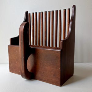 May include: A vintage wooden bread slicer with a dark brown finish. The slicer features a vertical slat back, a handle, and a rectangular base. The design is reminiscent of antique kitchen tools.