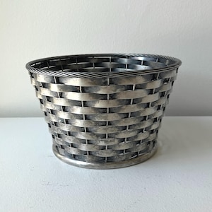 May include: A silver-toned metal basket with a woven design. The basket has a rounded base and a slightly flared top. The weave pattern is horizontal and vertical, creating a textured surface. The basket is likely used for storage or display.