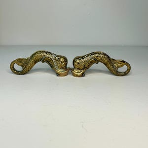 May include: A pair of matching brass drawer pulls shaped like stylized dolphins. Each piece features a curved body with detailed scale textures, a prominent fin, and a rounded head. The metal has a polished gold-toned finish with visible patina.