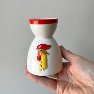 May include: A white ceramic egg cup with a red rim and a rooster design. The rooster has a red comb and wattle, and a yellow face and neck. The egg cup is held in a hand.