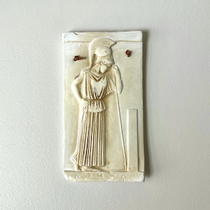 May include: Off-white rectangular relief sculpture of Athena, the Greek goddess of wisdom and warfare. The figure is depicted in classical attire, holding a staff. The word "ATHNA" is inscribed at the bottom. Two small holes are at the top.