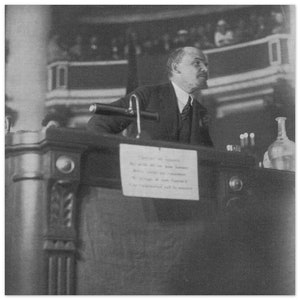 Op de afbeelding: Een man in pak spreekt achter een podium met een microfoon. Het podium heeft een bord met de tekst: "Voor de democratie. Maar wat bedoelen we met democratie? De oorsprong van het woord democratie. Het is essentieel voor het bestaan."