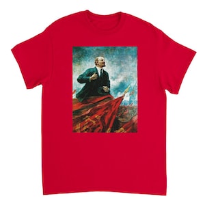 Peut inclure: T-shirt rouge avec une image vintage d'un homme en costume debout sur un drapeau rouge. L'homme tient un chapeau dans sa main droite et regarde vers la droite. L'image est sur fond de ciel bleu et blanc.