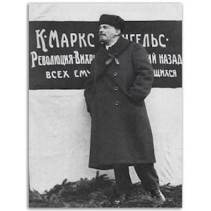 May include: A black and white photograph of a man in a long coat and fur-trimmed hat standing in front of a banner that reads "K.MARX ENGELS: REVOLUTION-WHIRLWIND OF ALL EMPIRES BACKWARDS"