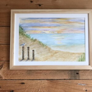 Peut inclure: Peinture à l'aquarelle d'une scène de plage dans un cadre en bois clair. L'œuvre représente un paysage côtier serein avec une plage de sable, l'océan et un ciel de coucher de soleil. Un petit oiseau est perché sur un poteau en bois au premier plan.