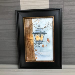 Pode incluir: Uma pintura em aquarela emoldurada de uma lanterna pendurada em um galho de árvore. A lanterna brilha com um brilho quente, com um pequeno pássaro empoleirado por perto. A pintura tem uma moldura preta com uma borda interna dourada.
