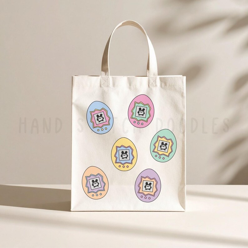 May include: A cream-colored canvas tote bag featuring a repeating pattern of six colorful egg-shaped digital pet devices. Each device is a different pastel color, including blue, pink, yellow, green, orange, and purple, with a small screen display.