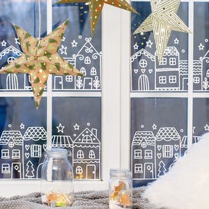 Puede incluir: Una ventana decorada con diseños de casas y estrellas blancas. Dos tarros de cristal con luces y elementos decorativos se encuentran sobre una superficie gris. Tres adornos de papel en forma de estrella con patrones festivos cuelgan.