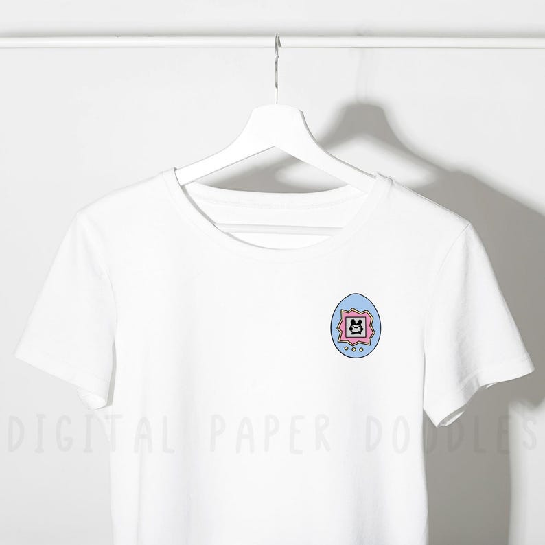 May include: A white short-sleeved t-shirt featuring a small graphic on the left chest. The graphic is a light blue and pink egg-shaped digital pet device with a screen displaying a black pixelated character and three small circular buttons below.