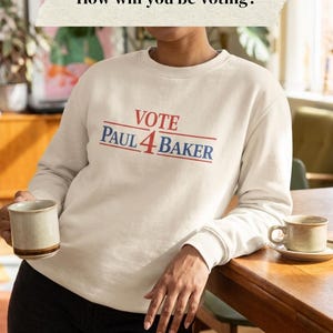 May include: A cream-coloured crewneck sweatshirt featuring the red and blue text VOTE PAUL 4 BAKER centred on the chest. The design includes horizontal red lines above and below the name. The text How will you be voting? appears above.