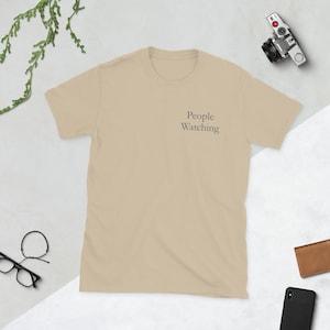 May include: A tan-colored t-shirt with the text "People Watching" printed in a dark gray font on the upper left chest area. The shirt is short-sleeved and has a crew neck.