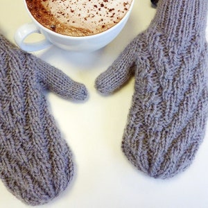 Könnte beinhalten: Ein Paar graue Strickhandschuhe mit einem Zopfmuster. Die Handschuhe liegen auf einer weißen Oberfläche.