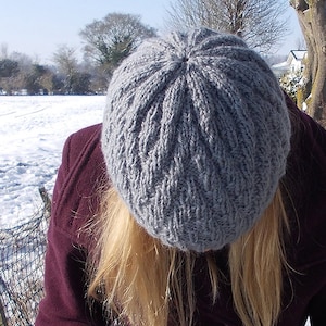 Puede incluir: Un gorro de punto gris con un patrón texturizado. El gorro lo lleva una persona con el pelo largo y rubio.