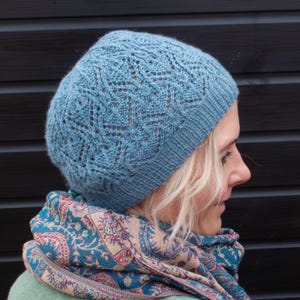 May include: A light blue knitted hat with a lacy pattern. The hat is worn by a person with blonde hair and a patterned scarf.
