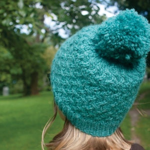 May include: A teal blue knitted hat with a fluffy pom-pom on top. The hat is made of a soft, textured yarn.