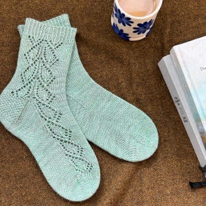 May include: A pair of light teal knitted socks with a decorative openwork pattern on the front. The socks are displayed on a brown surface, next to a floral-decorated cup and a book.