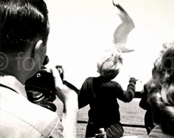 May include: A black and white photograph of three people watching a seagull fly overhead. The person on the left is holding a camera and taking a picture. The person in the middle is wearing a dark jacket and is looking up at the seagull. The person on the right is looking at the seagull with their hair blowing in the wind.