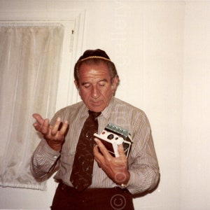 May include: A man wearing a striped shirt and a yarmulke holds a small, white, rectangular device in his right hand. He is gesturing with his left hand.
