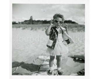 Può includere: Una bambina con un vestito bianco e una giacca a motivi bianco e nero sta in piedi su una spiaggia con uno sfondo bianco. Indossa occhiali da sole e ha i capelli tirati indietro.