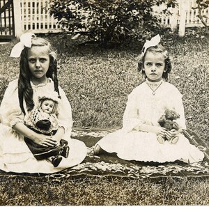 Deux filles effrayantes assises sur un tapis persan tenant une poupée et un ours en peluche dans un jardin, photo vintage, carte postale vintage