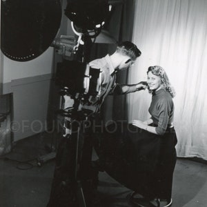 May include: A black and white photo of a man and a woman in a studio setting. The man is holding a camera and the woman is sitting on a stool. The man is looking at the woman and the woman is looking at the camera.
