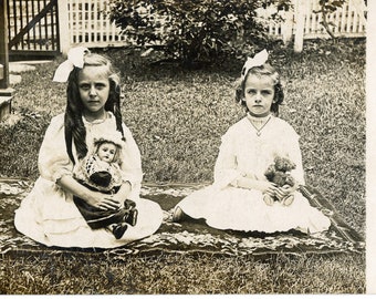 Dos chicas espeluznantes sentadas en la alfombra persa sosteniendo una muñeca y un oso de peluche en un patio trasero, foto vintage, postal vintage