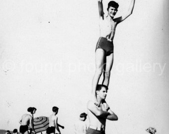 May include: Two people on a beach, one standing on the shoulders of the other. Both are wearing swimsuits. The person on top is raising their arms in the air. There are other people in the background.